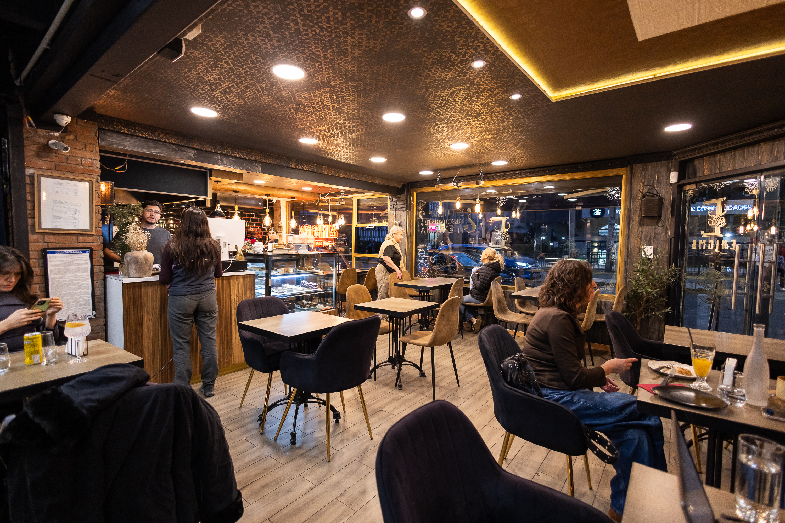 Salón de Enigma Café: mesas de madera, sillas de terciopelo azul, luz cálida y la barra al fondo