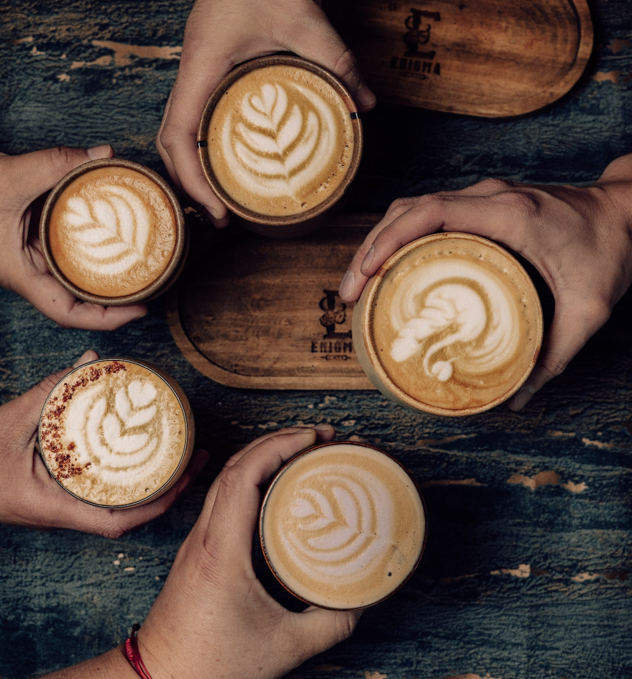 Cinco cafés con arte latte vistos desde arriba, sostenidos por varias manos sobre la mesa