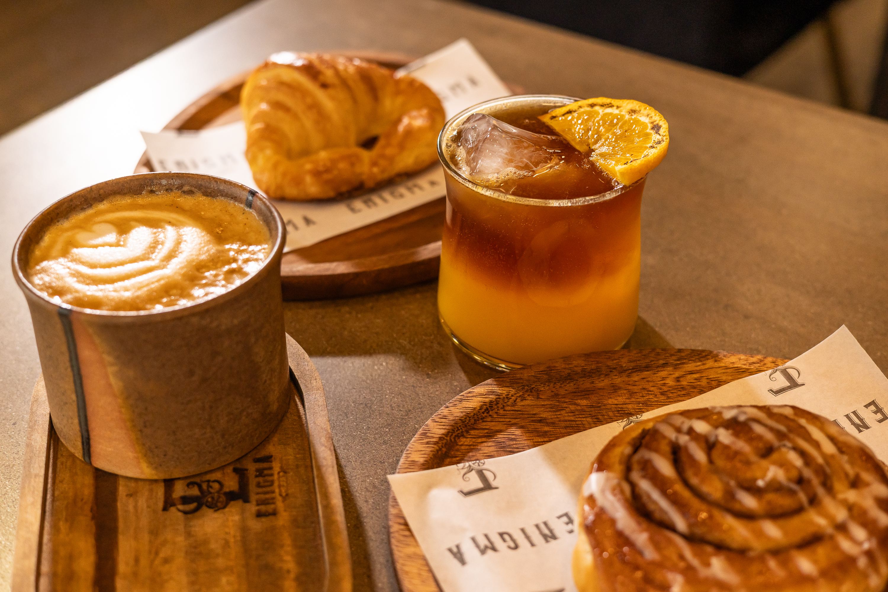 Café con leche, espresso tonic, croissant y rollo de canela sobre tablas Enigma